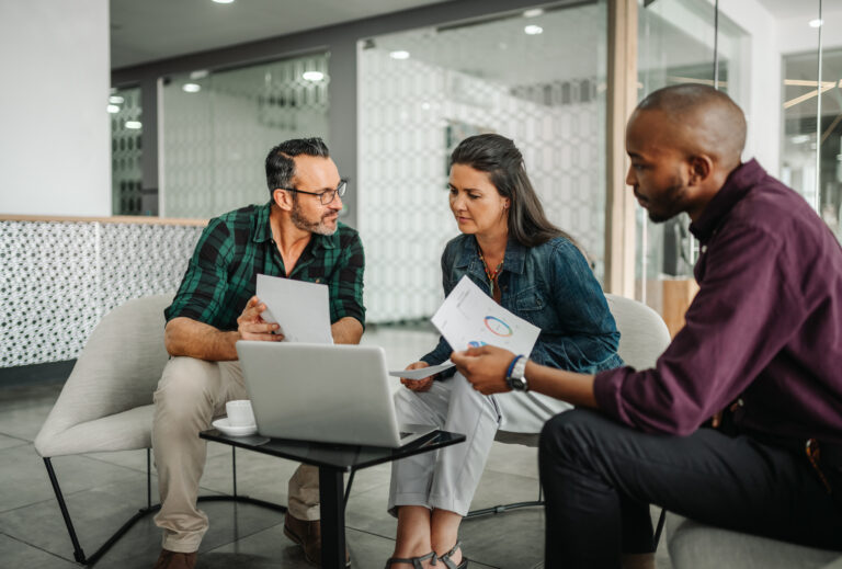 3 professional looking people holding papers and siting in a semi-circle Infront of laptops, appearing to be teaching each other.